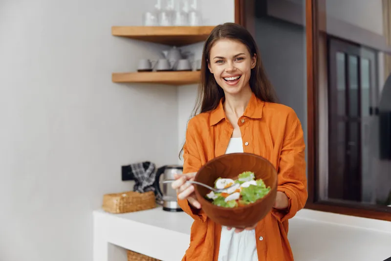 Счастливая молодая женщина в оранжевой рубашке демонстрирует вкусную миску с салатом в светлой кухне, что символизирует здоровое питание и энергичный образ жизни