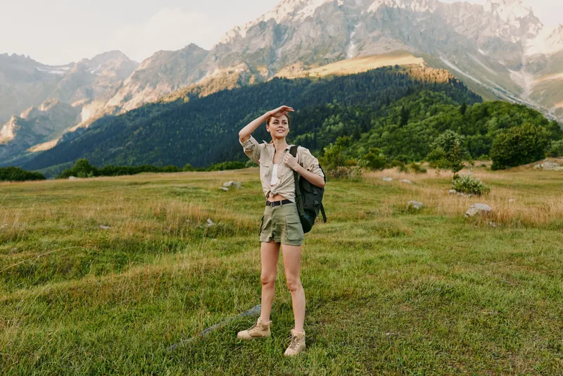 Woman with backpack standing in a grassy meadow near mountains, ready for adventure. Outdoor lifestyle scene with natural light and expansive landscape.