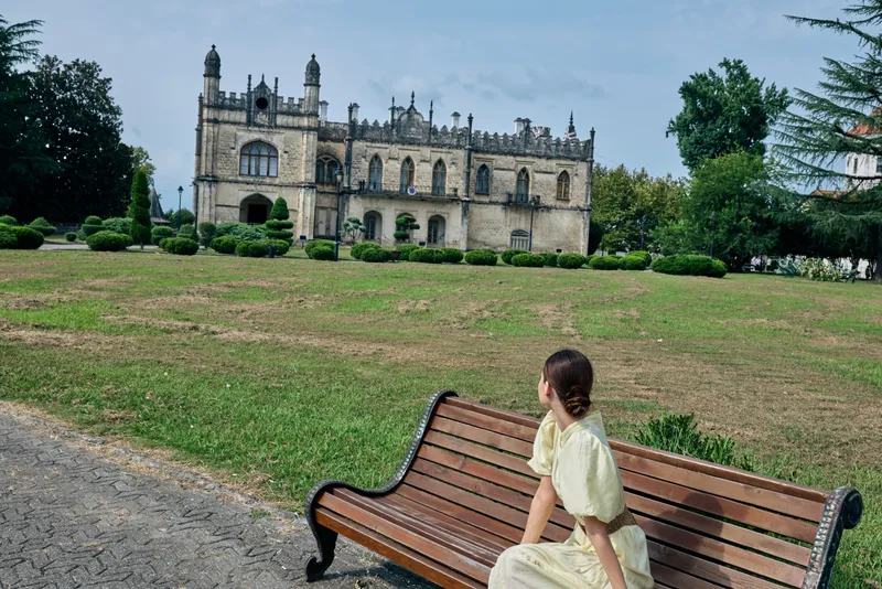 Young girl sits on a bench in a scenic park admiring a grand historic castle and expansive landscape, blending nature and heritage in a peaceful setting.