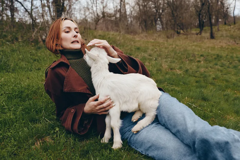 Woman lies on grass outdoors, gently pets a white goat kid, enjoying a calm lifestyle moment, casual clothing, peaceful park setting, close bond with animal