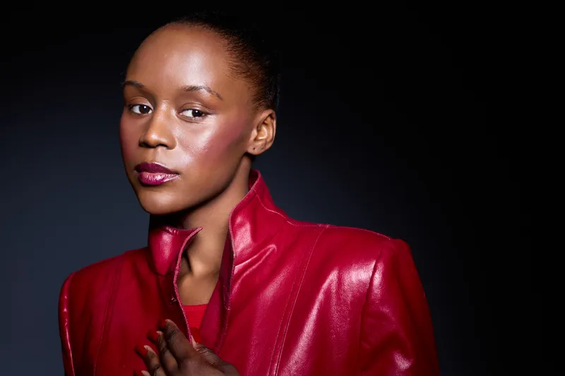 Close-up portrait of confident African woman with dark skin wearing red leather jacket. Studio shot with black background. Concept of fashion, beauty, and elegance with strong mood.