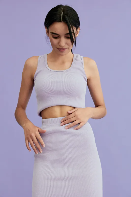 Young woman wearing a lilac crop top and matching skirt poses gracefully against a solid lavender background, showing style and calm confidence.