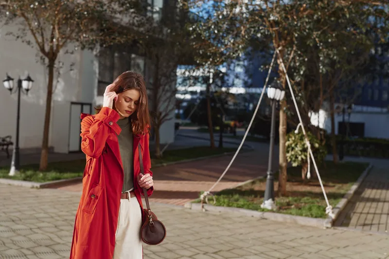A young woman in a red trench coat walks outdoors on a sunny day, holding her hair with one hand and carrying a round brown bag. Urban park setting with trees and lamp posts.