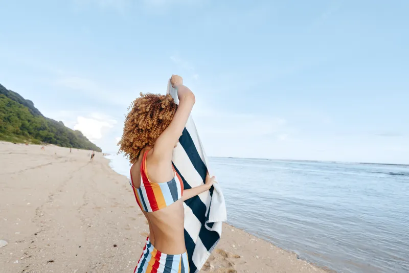 One person with curly hair wearing a striped swimsuit on a sandy beach, drying with a striped towel as waves roll in, capturing a relaxed summer mood.