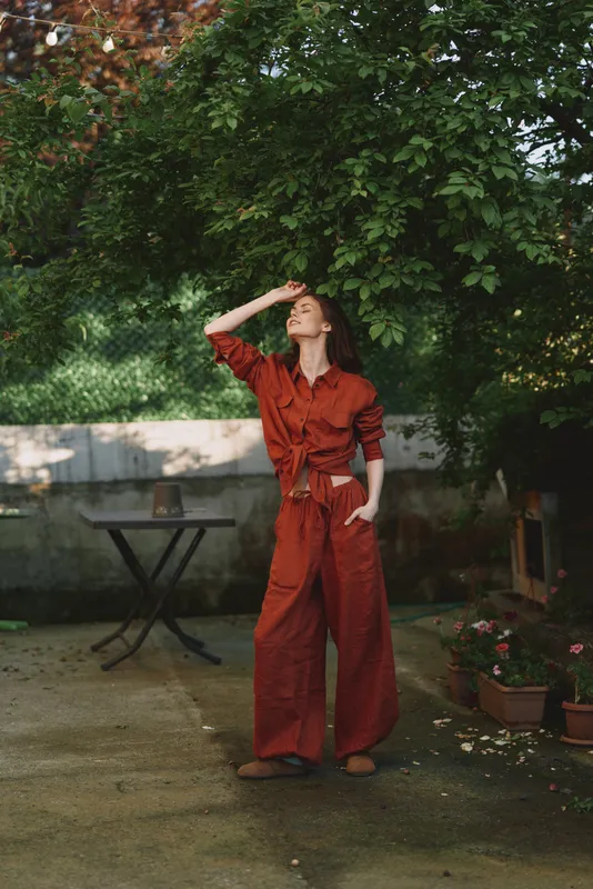 Smiling woman in a red jumpsuit stands outdoors under a leafy canopy, posing with one hand shading her eyes. Fashion lifestyle portrait with natural daylight and a garden backdrop