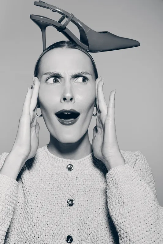 Surprised white woman with pale skin holds head with hands and balances high heel shoe on head in studio.