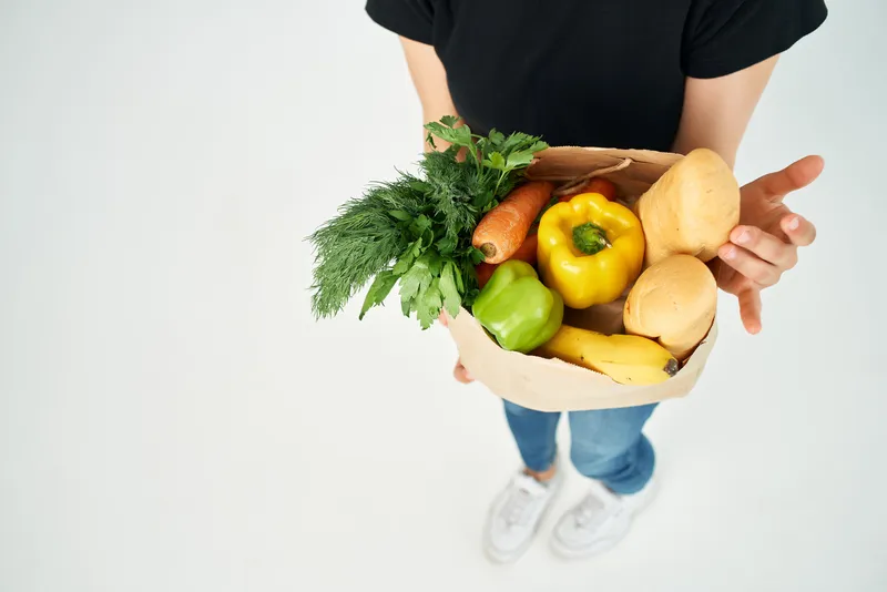 В руке человека находится пакет с продуктами, наполненный овощами, что символизирует доставку здоровой пищи