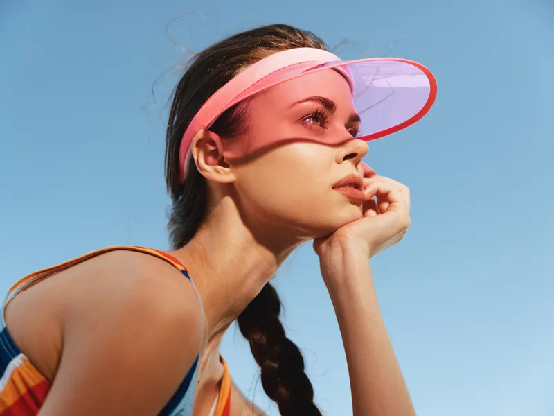 Portrait of a young woman wearing a pink visor outdoors, sunlight highlights her thoughtful expression as she rests her chin on her hand against a clear blue sky.