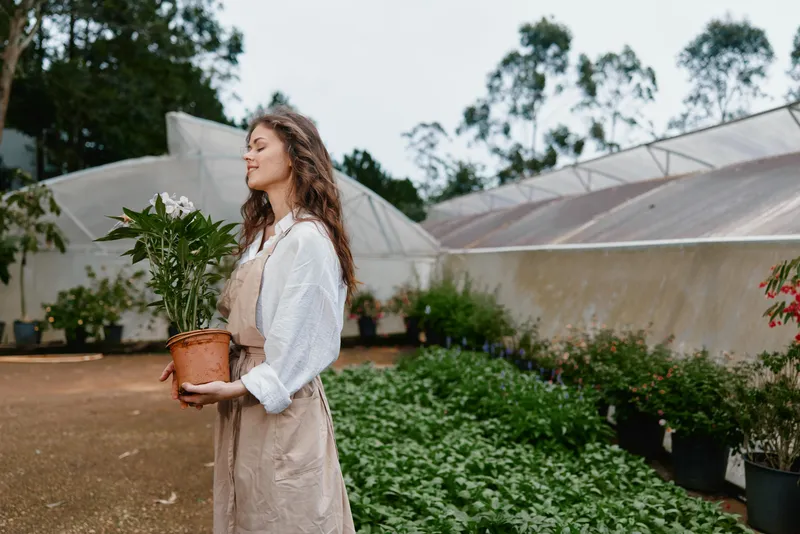 Young woman holding a potted plant in a greenhouse, showcasing eco friendly living and gardening passion with natural light illuminating her smile