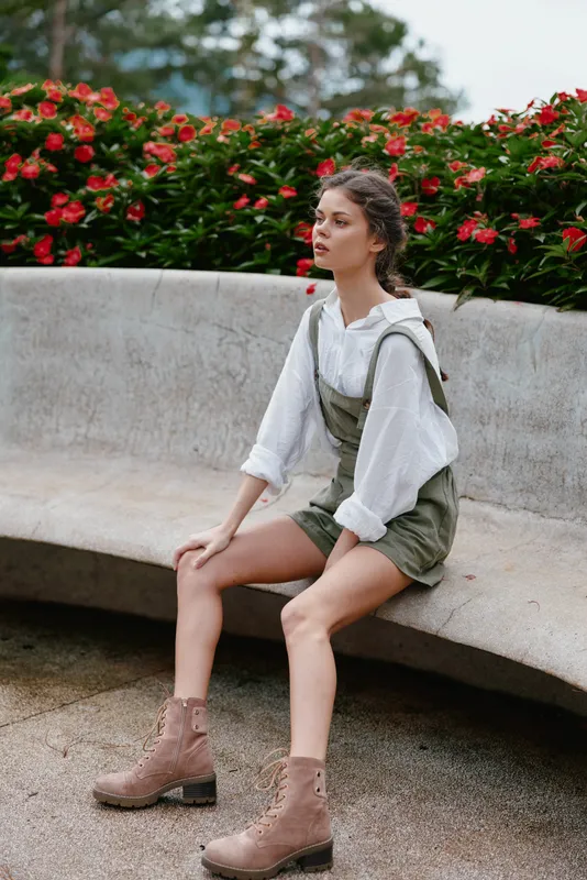 Stylish young woman in casual summer outfit sitting on a bench in a park surrounded by blooming flowers, showcasing a relaxed and natural vibe