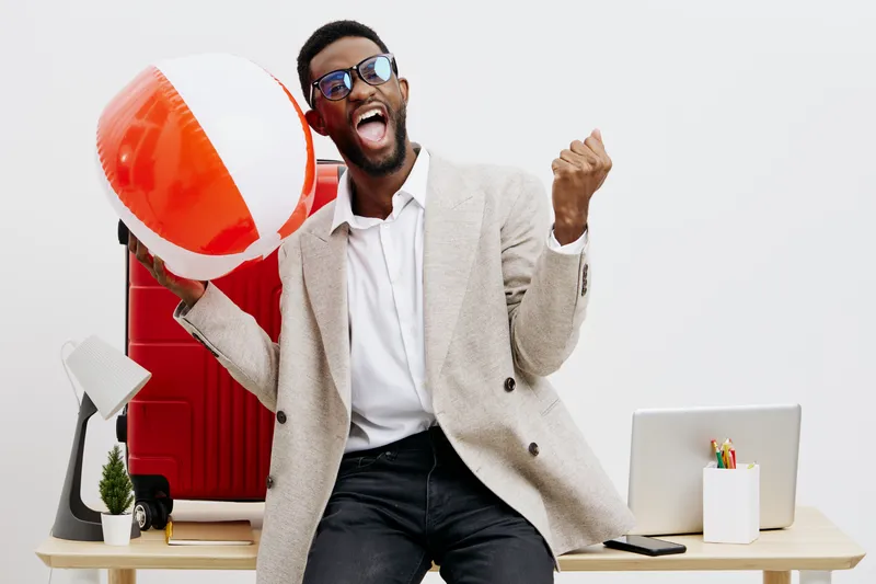 Cheerful young man with dark skin wearing sunglasses, beige blazer, and white shirt, holding colorful beach ball, celebrating happiness in bright studio setting with white background. People lifestyle