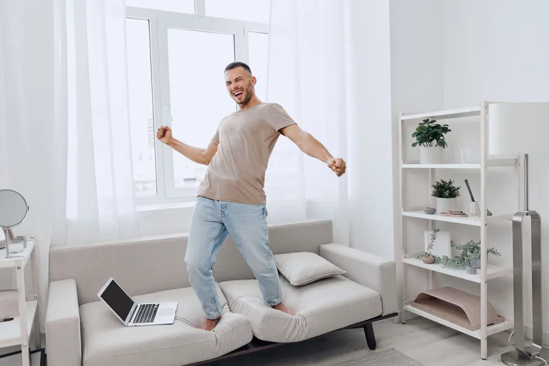 Joyful man celebrating success while standing on a couch in spacious, bright living room. This trendy scene captures modern home-office vibes effortlessly.
