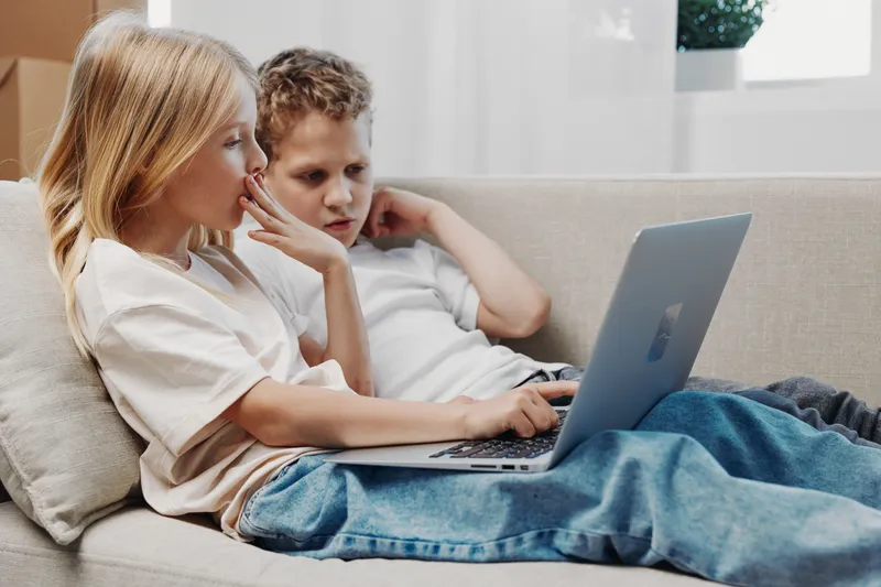 Two children sitting on a sofa using a laptop together with focused expressions. Cozy home interior with natural light, concept of technology, learning, and digital childhood experience.
