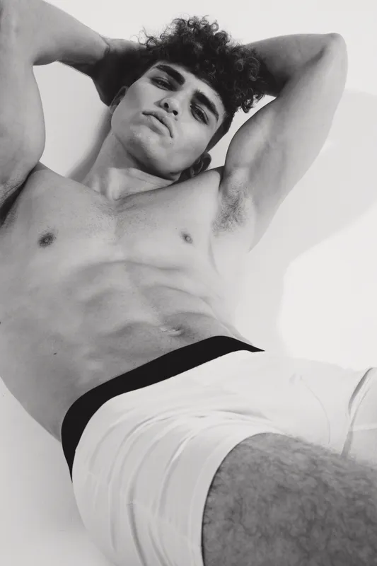 Young man with curly hair posing shirtless in white underwear, showing muscular torso and confident expression in black and white studio portrait with soft lighting.