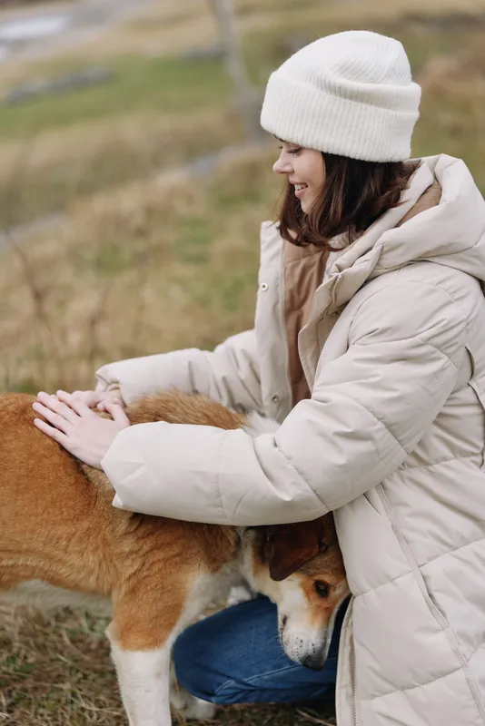 woman, dog, hug, outdoor, coat, beanie in a chilly field, a smiling woman gently pats a dog beside open grass and hills, conveying warmth, care and companionship in nature