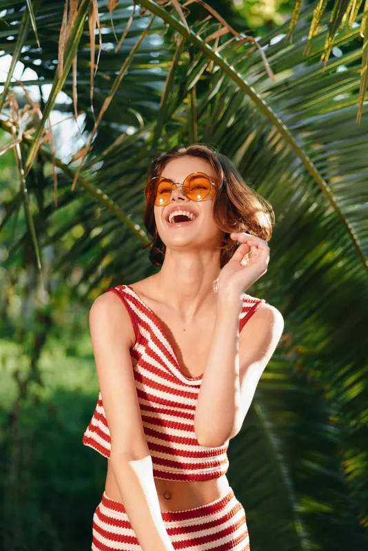 A cheerful woman wearing a red striped bikini laughs in a sunny outdoor setting, surrounded by lush tropical greenery, radiating a vibrant, carefree mood.
