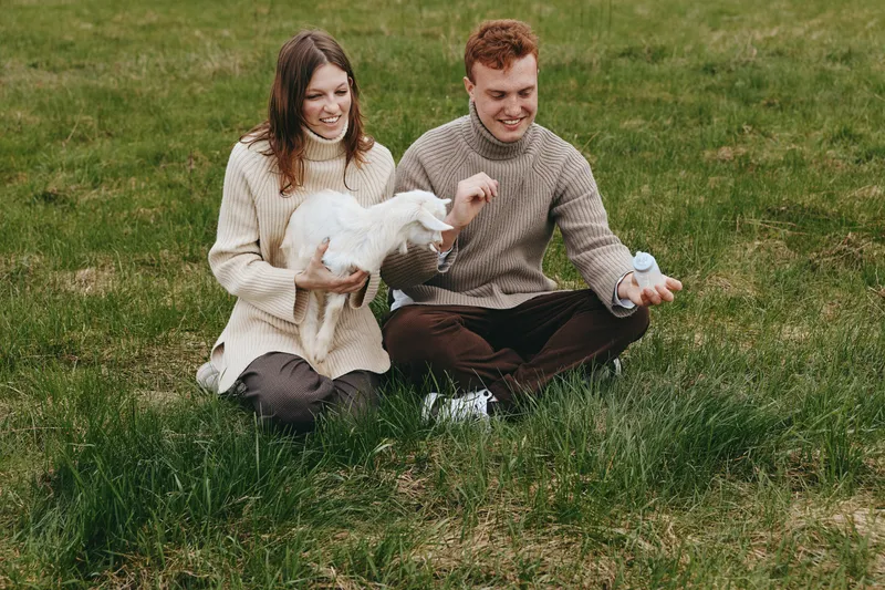 Young couple sitting on grass in casual sweaters enjoys spending time with a baby goat in a natural outdoor setting during a relaxed lifestyle moment.