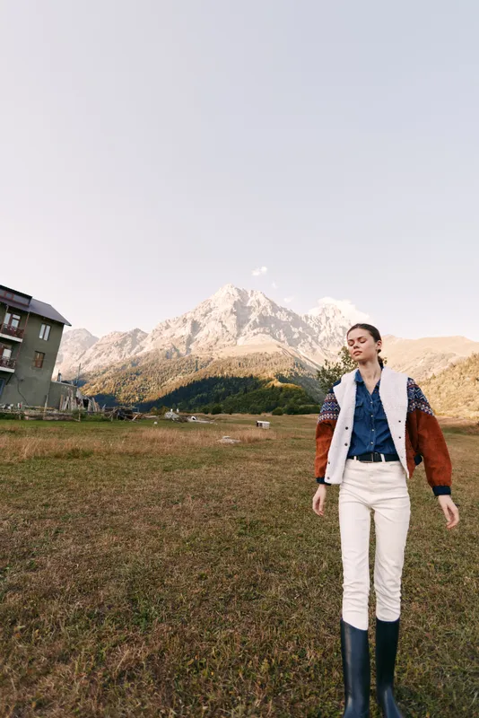 Woman in countryside field with towering mountains behind, fashion portrait of a model wearing denim jacket and boots, walking in a meadow at golden light and calm outdoor lifestyle.