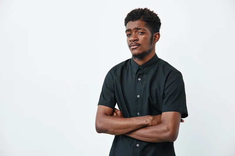 Young black man with dark skin and curly hair wearing black shirt standing with crossed arms on white background. Portrait of confident african american male with serious expression.