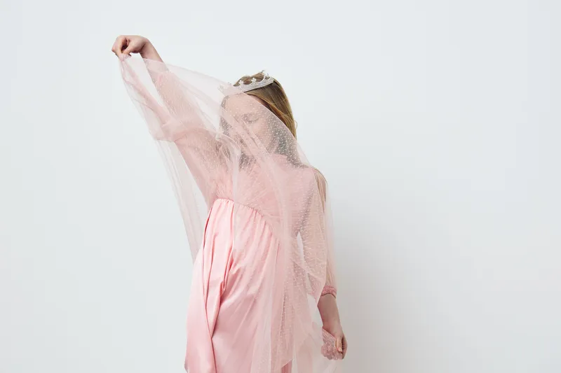 A young girl wearing a pink dress and a crown, playfully covering her face with a sheer veil, exuding joy and innocence against a minimalistic white background