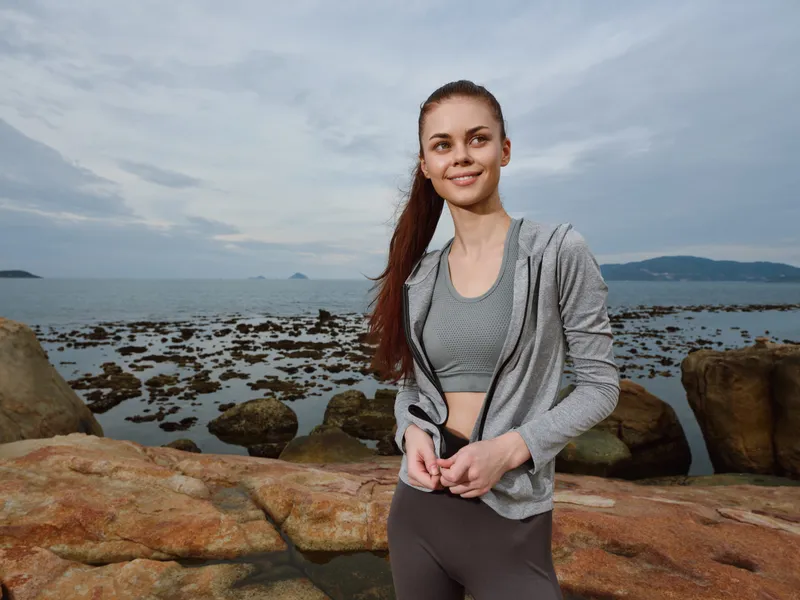 Fit woman in sportswear smiling by the sea, embodying health and vitality, wearing a gray jacket and athletic leggings, reflecting a lifestyle of wellness and fitness