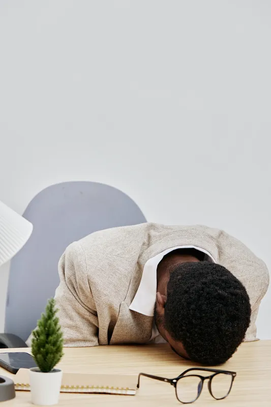 Professional photograph of a man with dark skin and curly black hair, sitting at a desk with his head down on the table, wearing a beige sweater and white shirt, with glasses, in a modern office setti