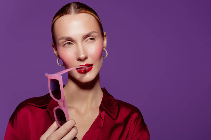 Stylish woman in red blouse holds pink sunglasses near lips, posing on purple background. Concept of fashion, beauty, accessories, and confident female model with trendy look.
