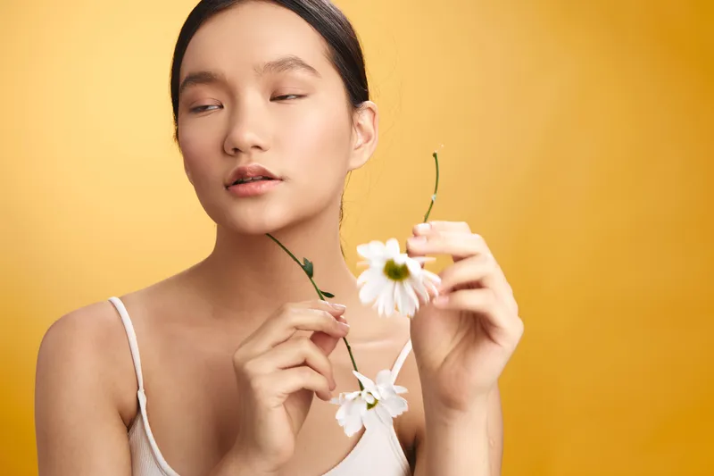 asian woman adjusts delicate petals in a soft portrait against a warm yellow backdrop, conveying calm beauty and gentle focus in a minimal, serene setting