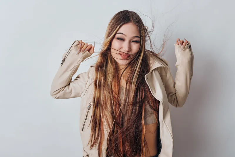 Young woman with long hair wearing beige jacket posing with playful expression on light background. Female model showing fun and carefree mood with natural look and casual style.
