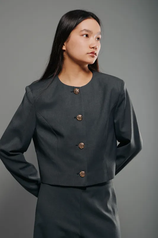 Young woman in dark gray suit with gold buttons posing looking sideways against plain background. Fashion model wearing formal outfit with long hair and serious expression.