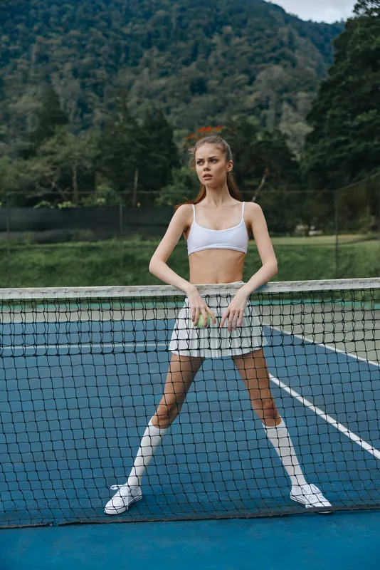 Young female athlete in a white crop top and skirt posing confidently on a tennis court, showcasing athleticism and style against a natural backdrop