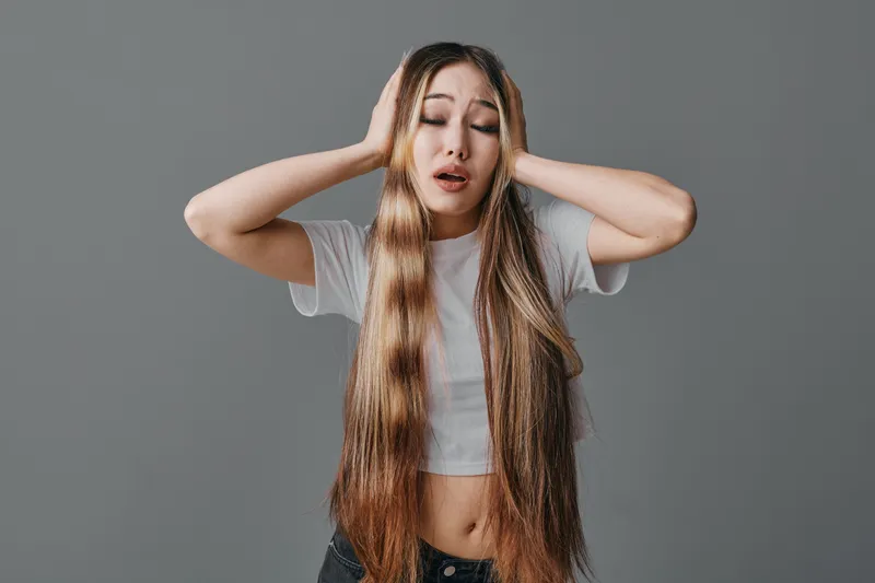 Young woman with long hair holds head with hands and closes eyes for headache or stress isolated on gray background in studio. Female model expressing pain and discomfort.