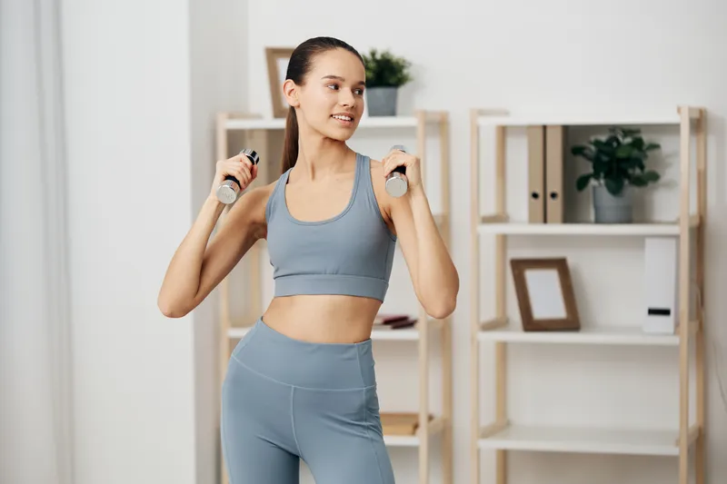 Woman fitness, sport and workout scene featuring a fit woman in a blue sports set using dumbbells for arms training in a bright home gym. Clear focus on form, balance, and motivation for health and
