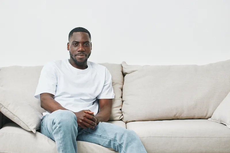 Young African American man sitting on a beige sofa, wearing a white t shirt and blue jeans, displaying a friendly expression in a minimalist living room