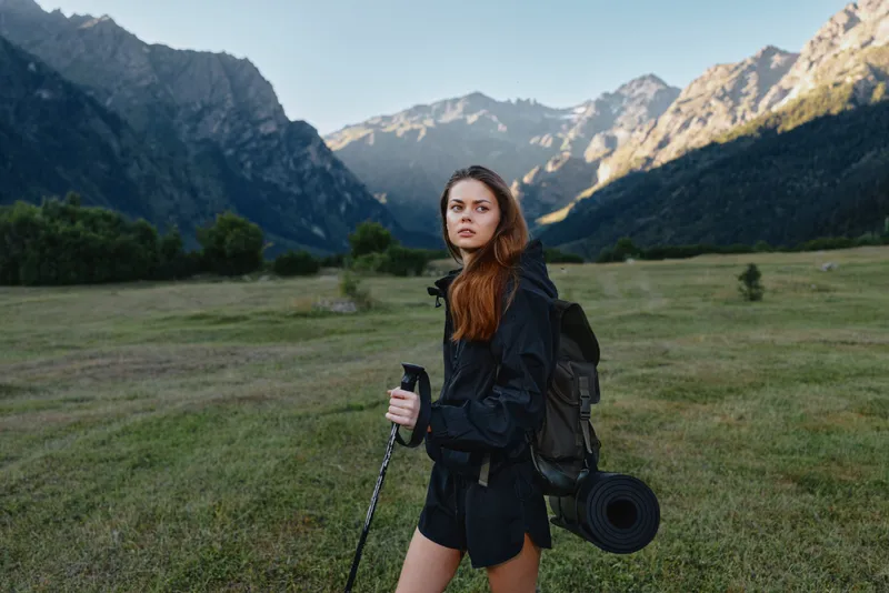 A lone female traveler stands in a grassy meadow surrounded by towering mountains. She wears a backpack and carries a hiking pole, prepared for a scenic outdoor adventure.