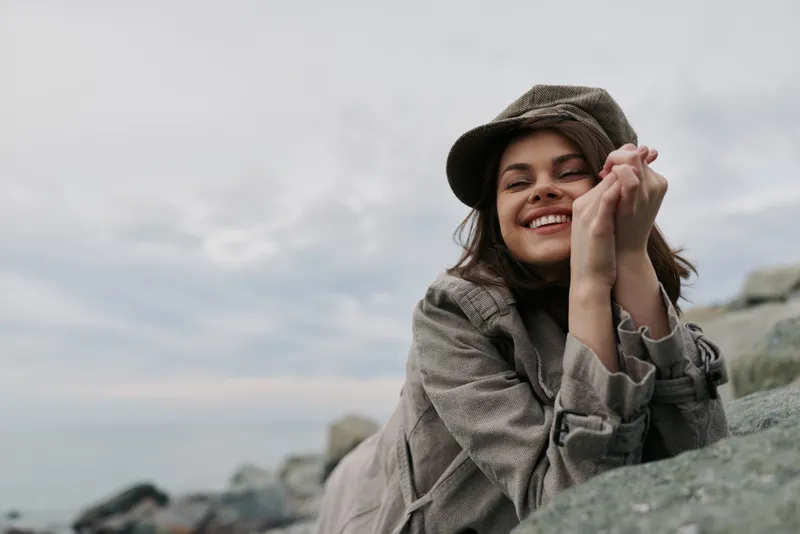 Smiling young woman in casual clothing and hat rests on rocks near calm sea under cloudy sky. Natural outdoor scene with relaxed mood and soft natural light, lifestyle and happiness concept.