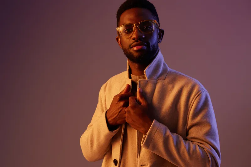 Stylish young Black man wearing trendy glasses and a fashionable coat, posing confidently against a colorful background, evoking a sense of modern fashion and self expression