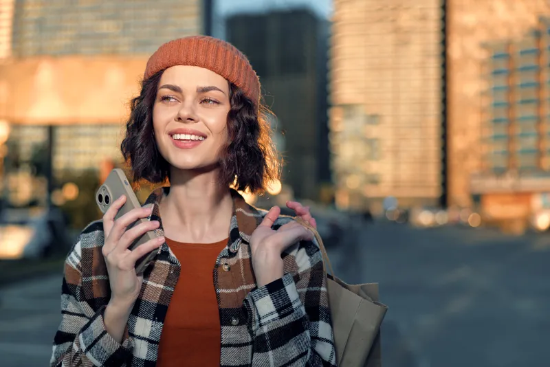 Young woman with smartphone smiling in an urban street at golden hour, candid lifestyle portrait that captures authenticity, joyful mood, warm glow and modern city living.