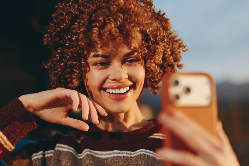 Woman lifestyle portrait with curly hair, bright smile, holding phone for a casual selfie, wearing a warm sweater outdoors, soft bokeh mountains background, communication and joy