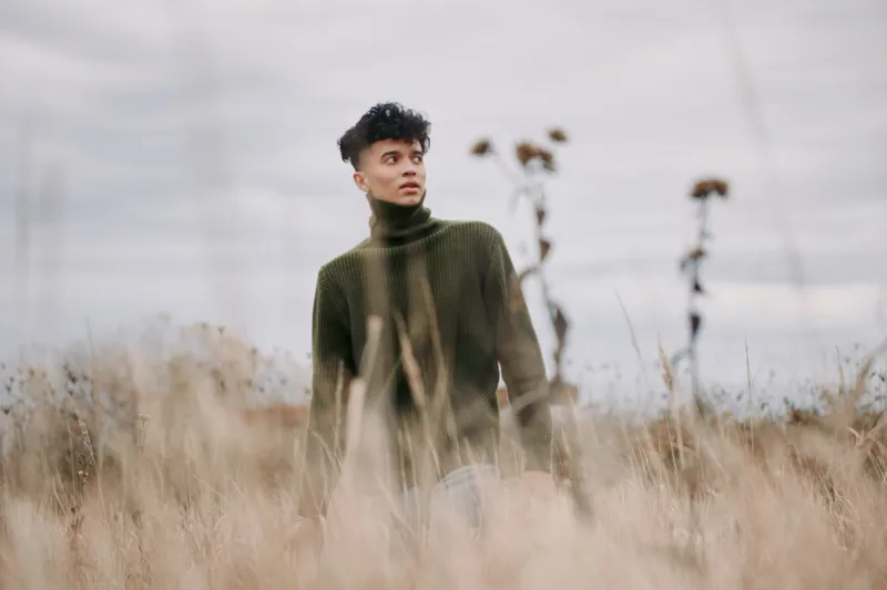 A young man in a green turtleneck stands amid tall dry grass, shot with natural light and candid framing to convey authenticity and a genuine, unposed portrait mood.