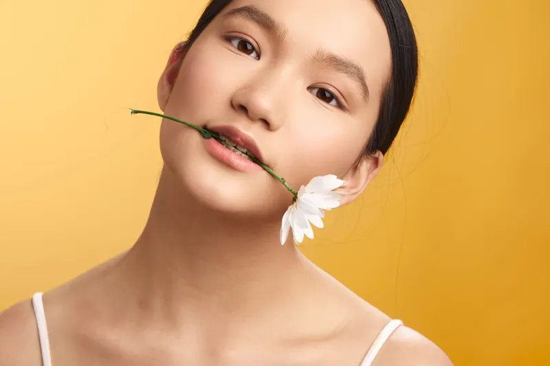 asian woman poses with a flower between her lips against a warm yellow backdrop, presenting a serene, stylish portrait that conveys natural beauty and gentle charm.