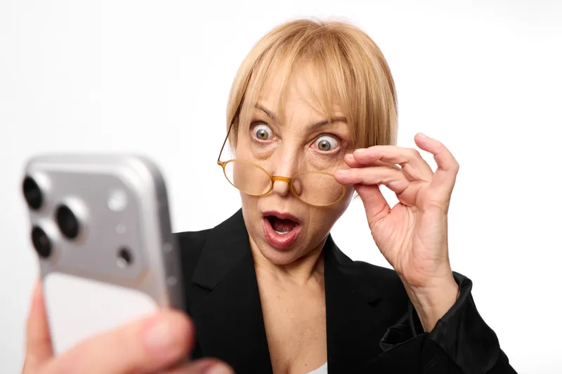 Woman businesswoman looks surprised and shocked, holding phone and glasses, wide eyes open mouth, stress and anxiety from news communication in office, workplace panic