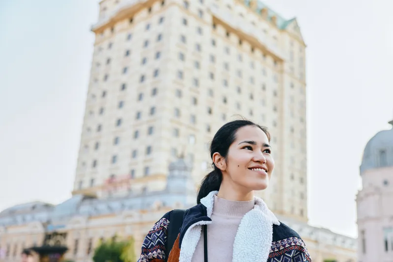 Woman smiling outdoors in city surroundings near a tall building, travel fashion, urban scenery, daylight, casual layers, confident pose, sightseeing mood, modern architecture