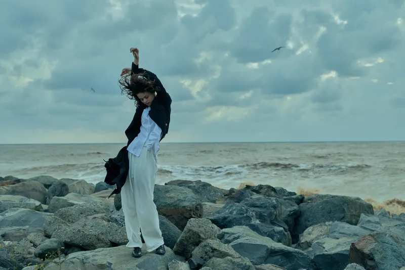 Woman standing on a rocky shoreline, white dress and dark jacket fluttering in the wind, vast sea and cloudy horizon behind, dramatic coastal fashion and outdoor mood, serene yet powerful scene