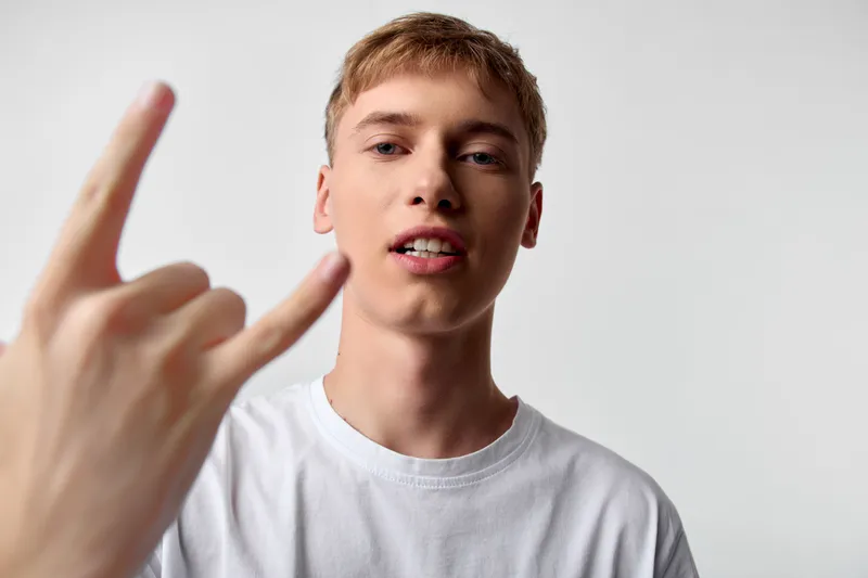 Rock on gesture from a young man in a white t shirt, close up framing with hand in foreground, casual style and confident expression in a clean studio setting