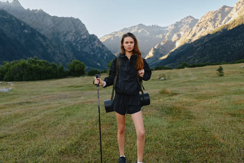 A confident hiker stands in a spacious valley under towering peaks, holding trekking poles. She wears outdoor gear and projects resilience, curiosity, and a ready spirit for adventure.