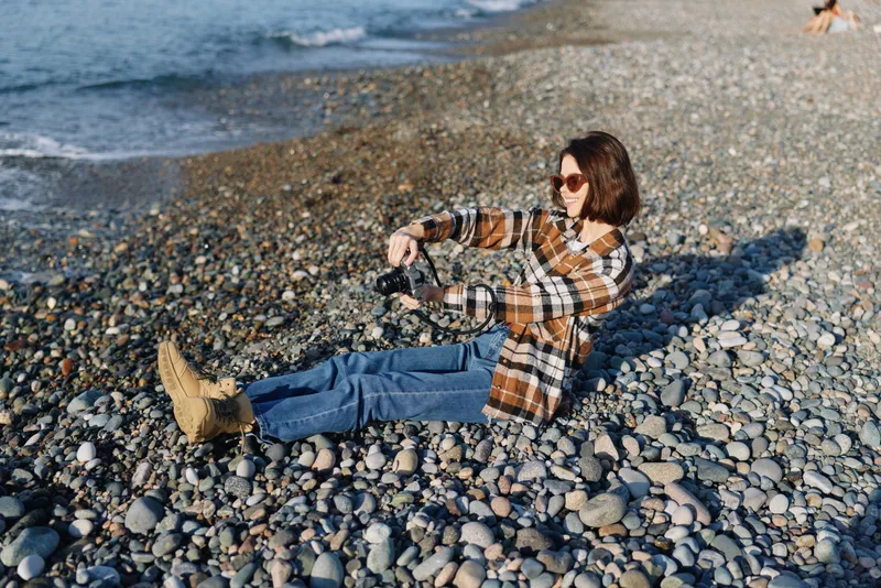 Casual photographer lying on pebbled beach with camera, enjoying sunny day by the sea, plaid shirt, jeans, boots, sunglasses, capturing coastal scenery and relaxed outdoor mood.