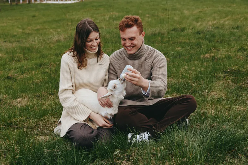 Couple outdoors feed a goat kid with a bottle, smiling and cozy in sweaters on a grassy day, lifestyle moment captured with warmth, care, and natural light.