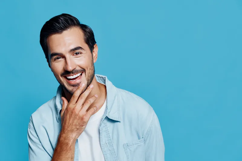 Smiling man in light denim shirt touching his chin, looking at the camera with a cheerful expression against a vibrant blue background. A portrait filled with positivity.