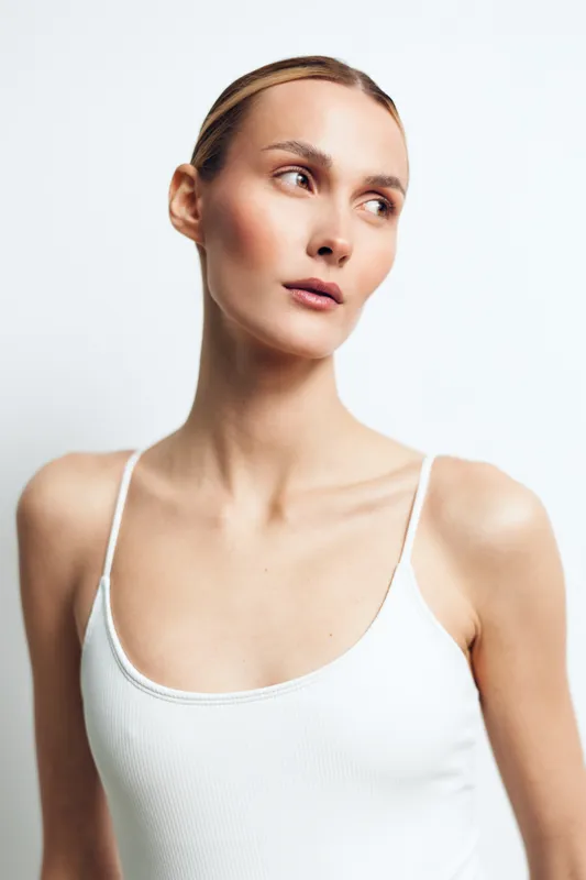 Young woman with fair skin wearing white tank top looking sideways against plain white background, natural light, minimal makeup, smooth skin, clean and simple style